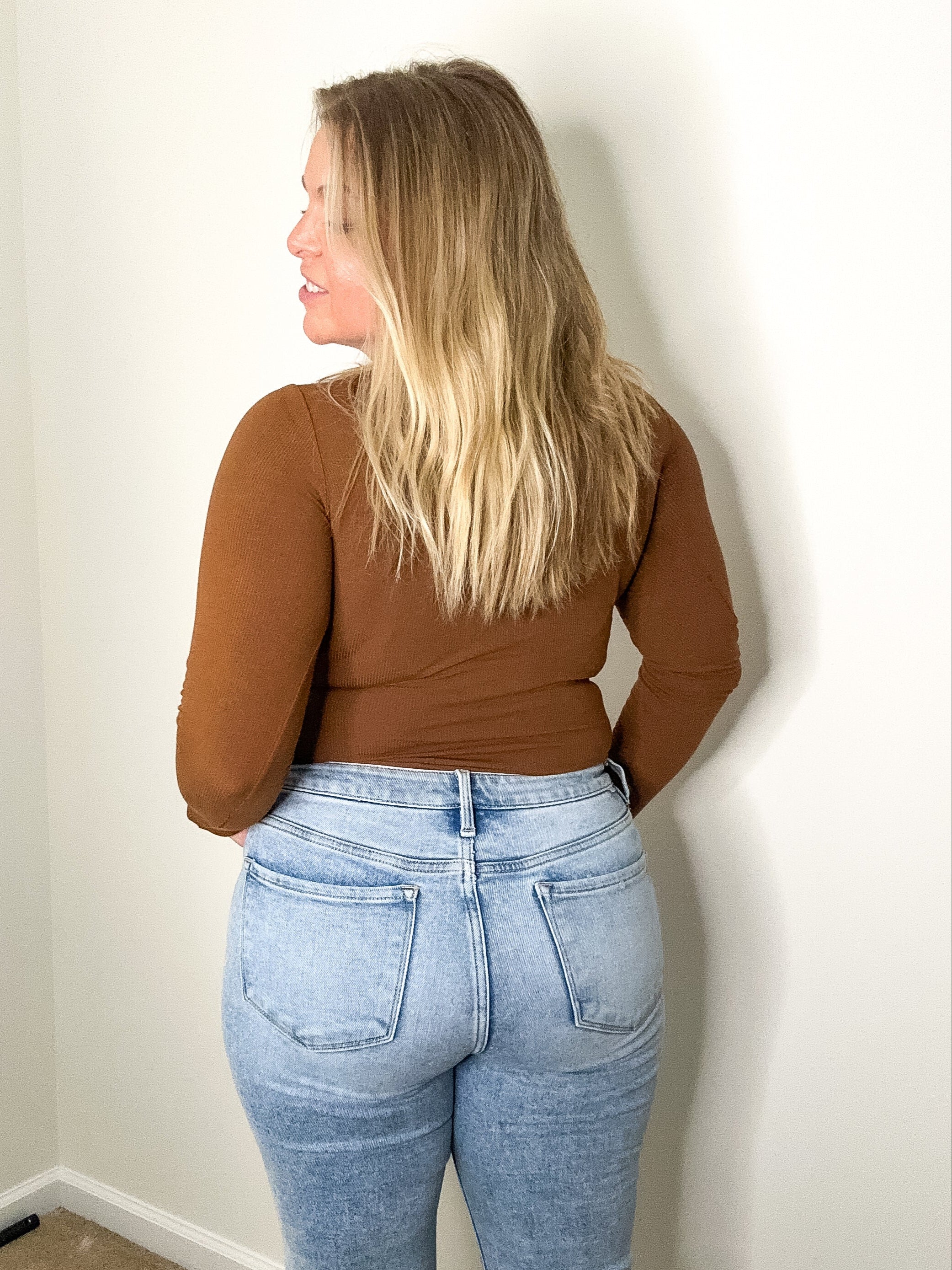 Person wearing a brown top and blue jeans against a plain wall.