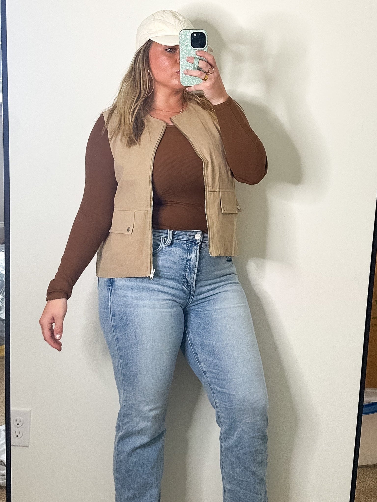 Person taking a mirror selfie wearing a brown top, beige vest, and blue jeans in a room with a large mirror.