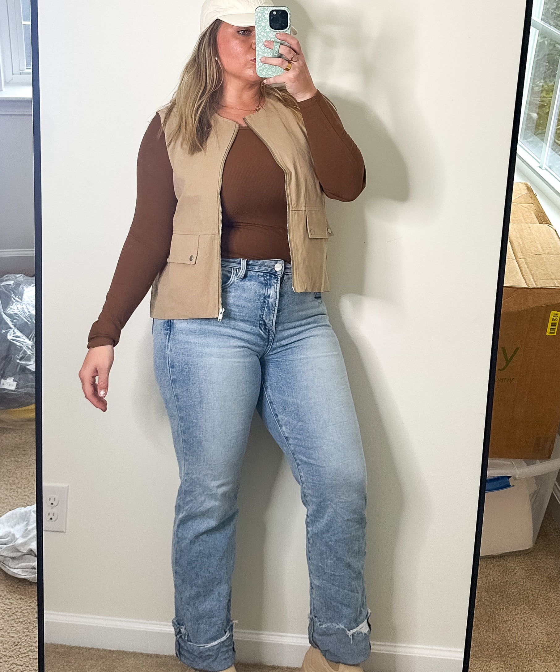 Person taking a mirror selfie wearing a brown top, beige vest, and blue jeans in a room with a large mirror.