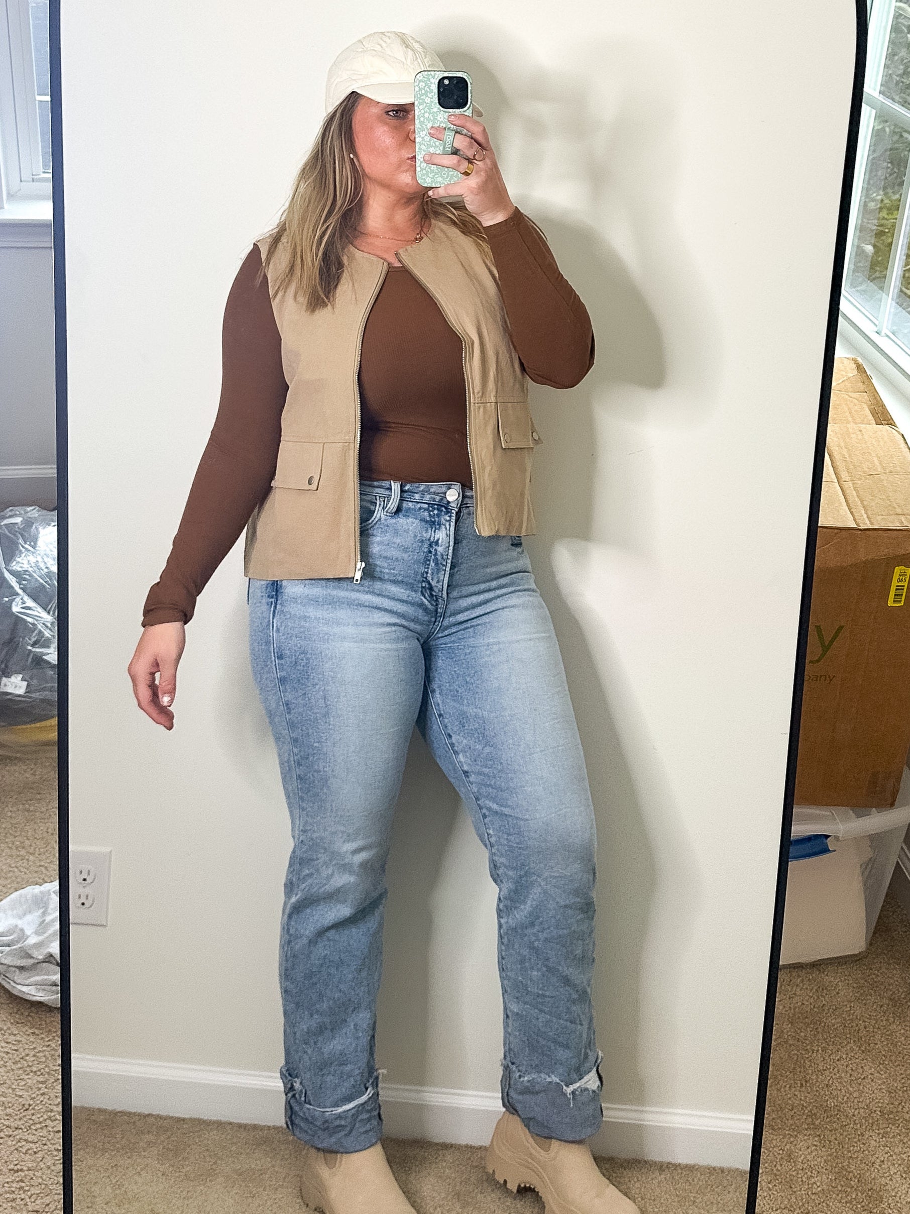 Person taking a mirror selfie wearing a brown top, beige vest, and blue jeans in a room with a large mirror.