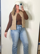 Person taking a mirror selfie wearing a brown top, beige vest, and blue jeans in a room with a large mirror.