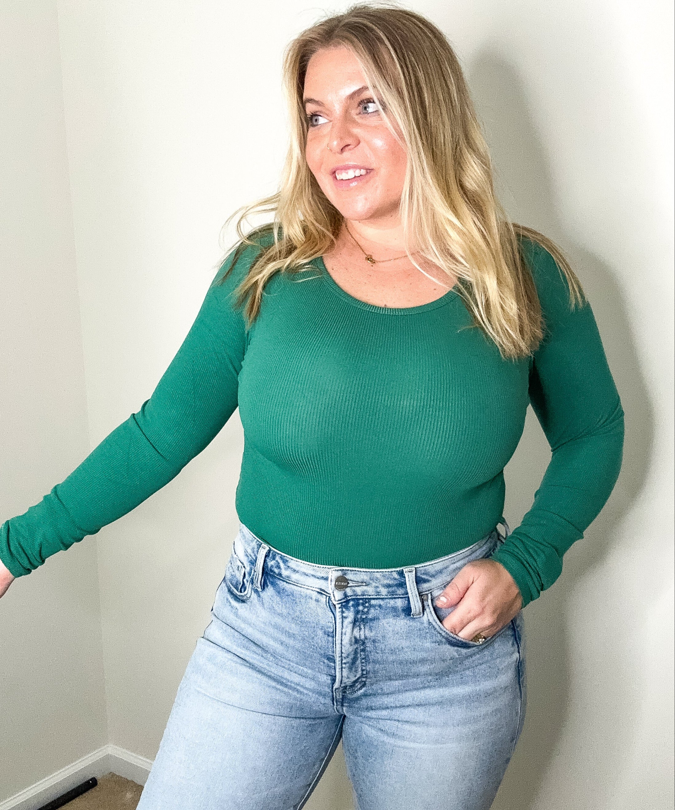 Woman wearing a green long-sleeve top and blue jeans against a plain wall.