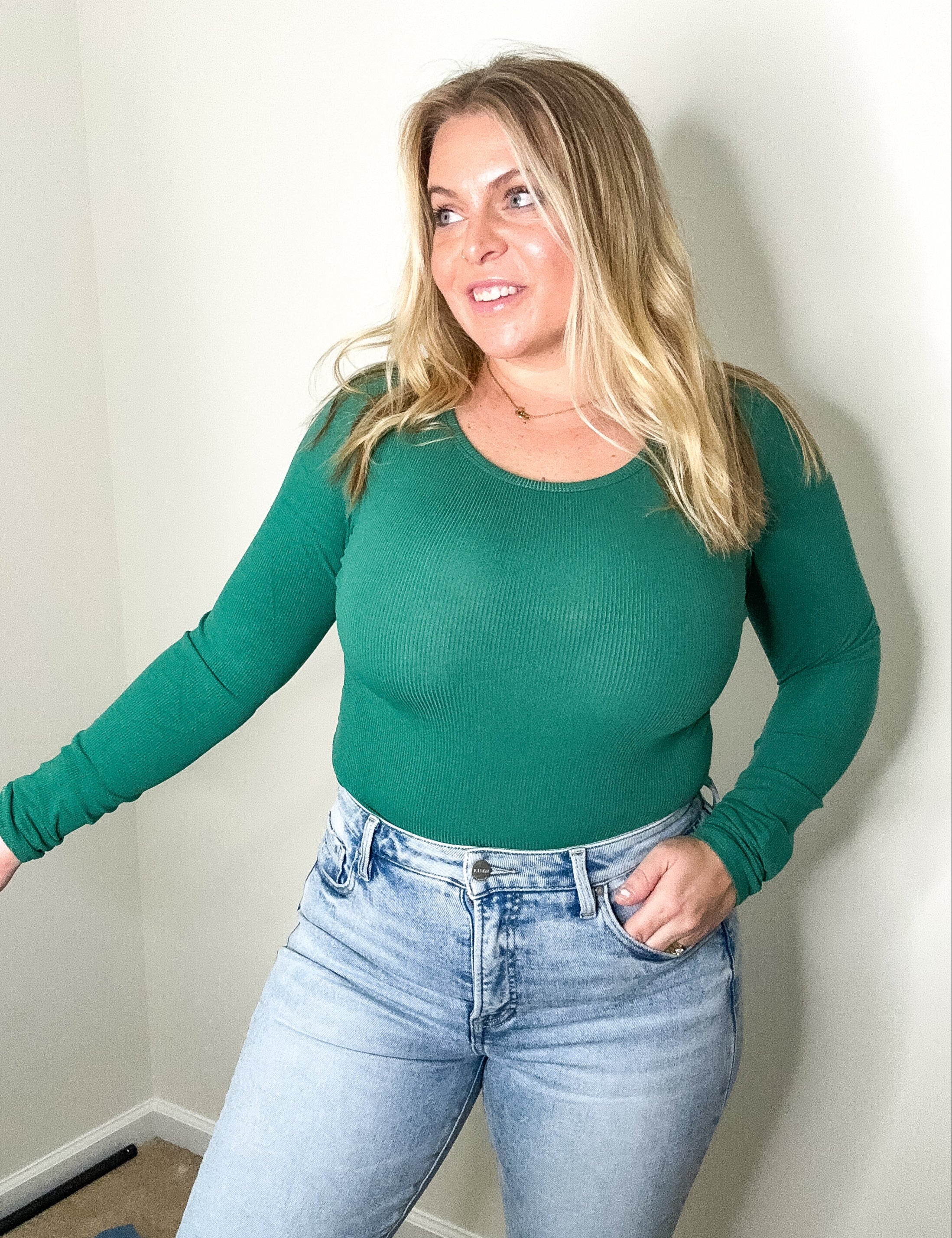 Woman wearing a green long-sleeve top and blue jeans against a plain wall.
