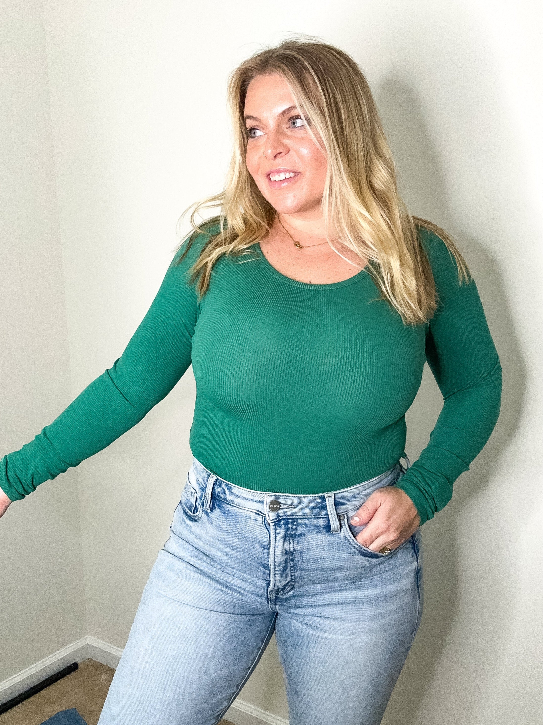 Woman wearing a green long-sleeve top and blue jeans against a plain wall.