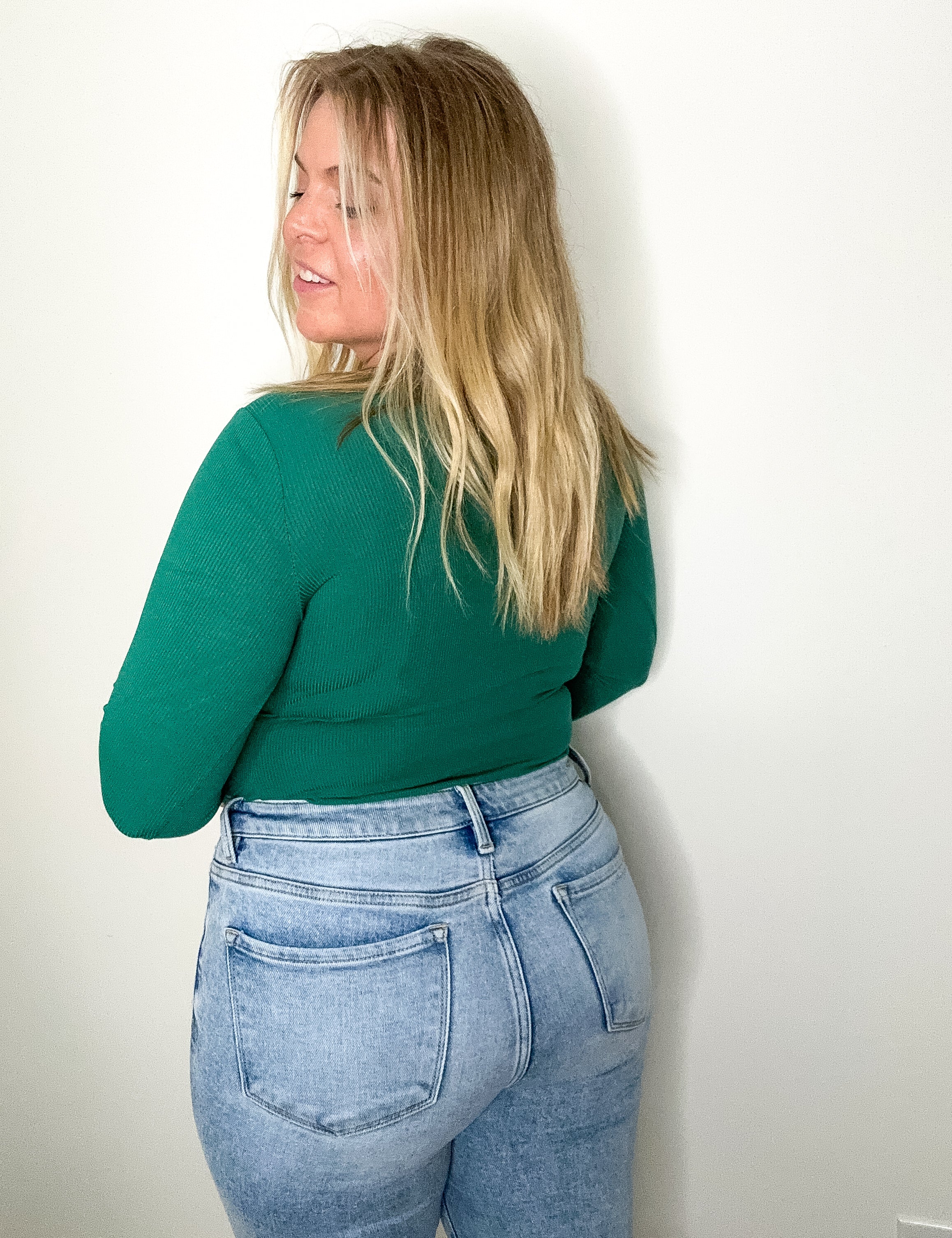 Person wearing a green top and light blue jeans against a white wall