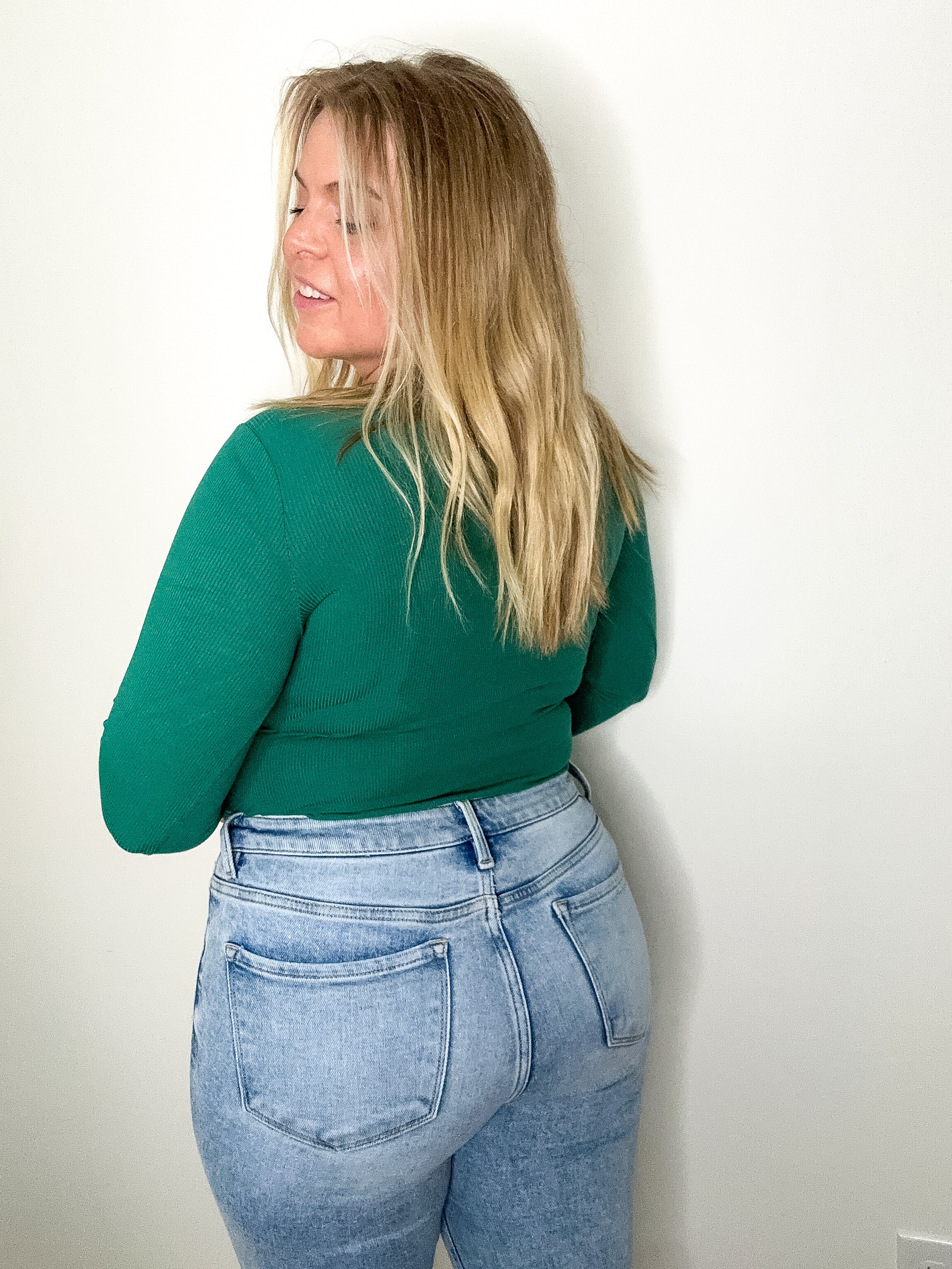 Person wearing a green top and light blue jeans against a white wall
