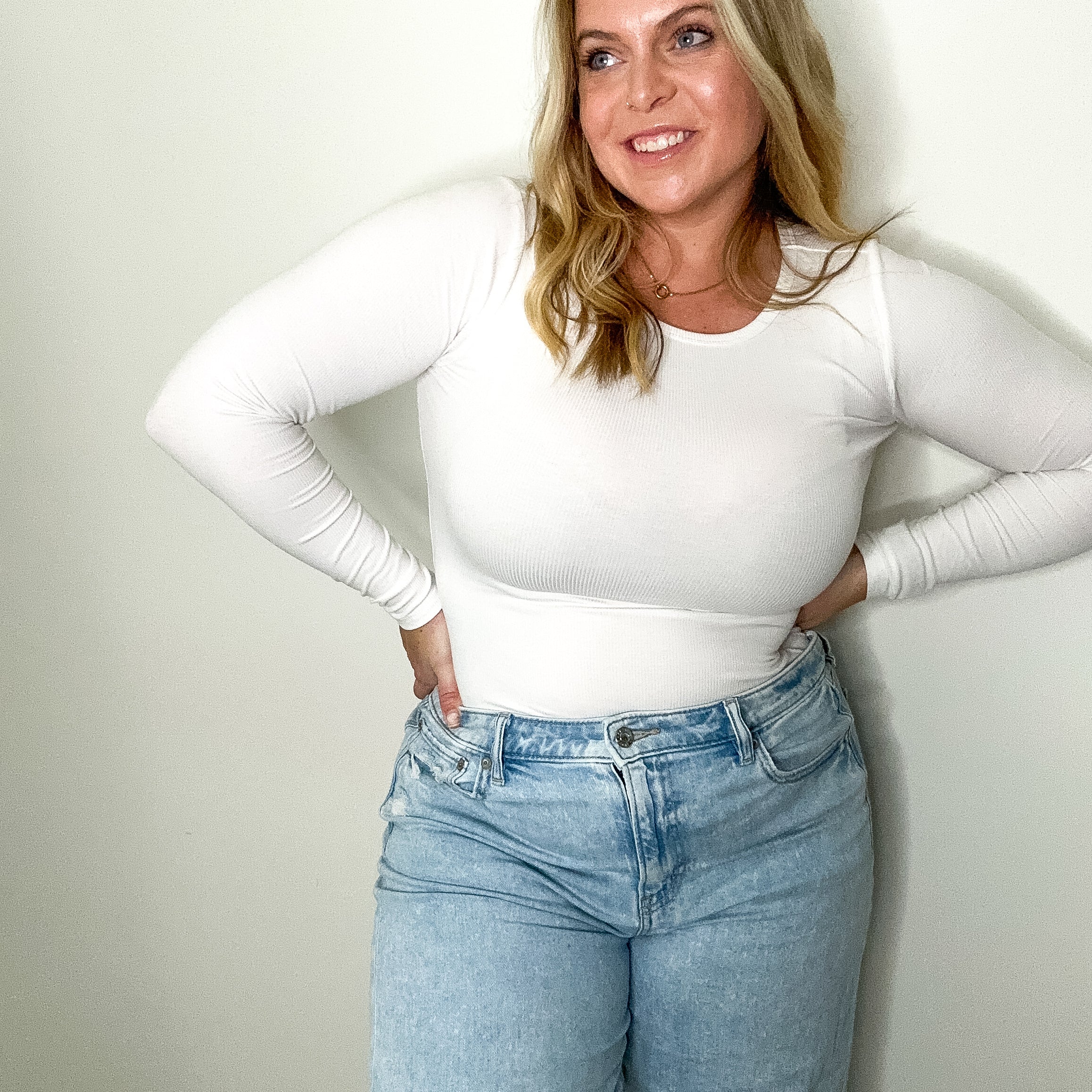 Woman wearing a white long-sleeve top and blue jeans against a plain wall.