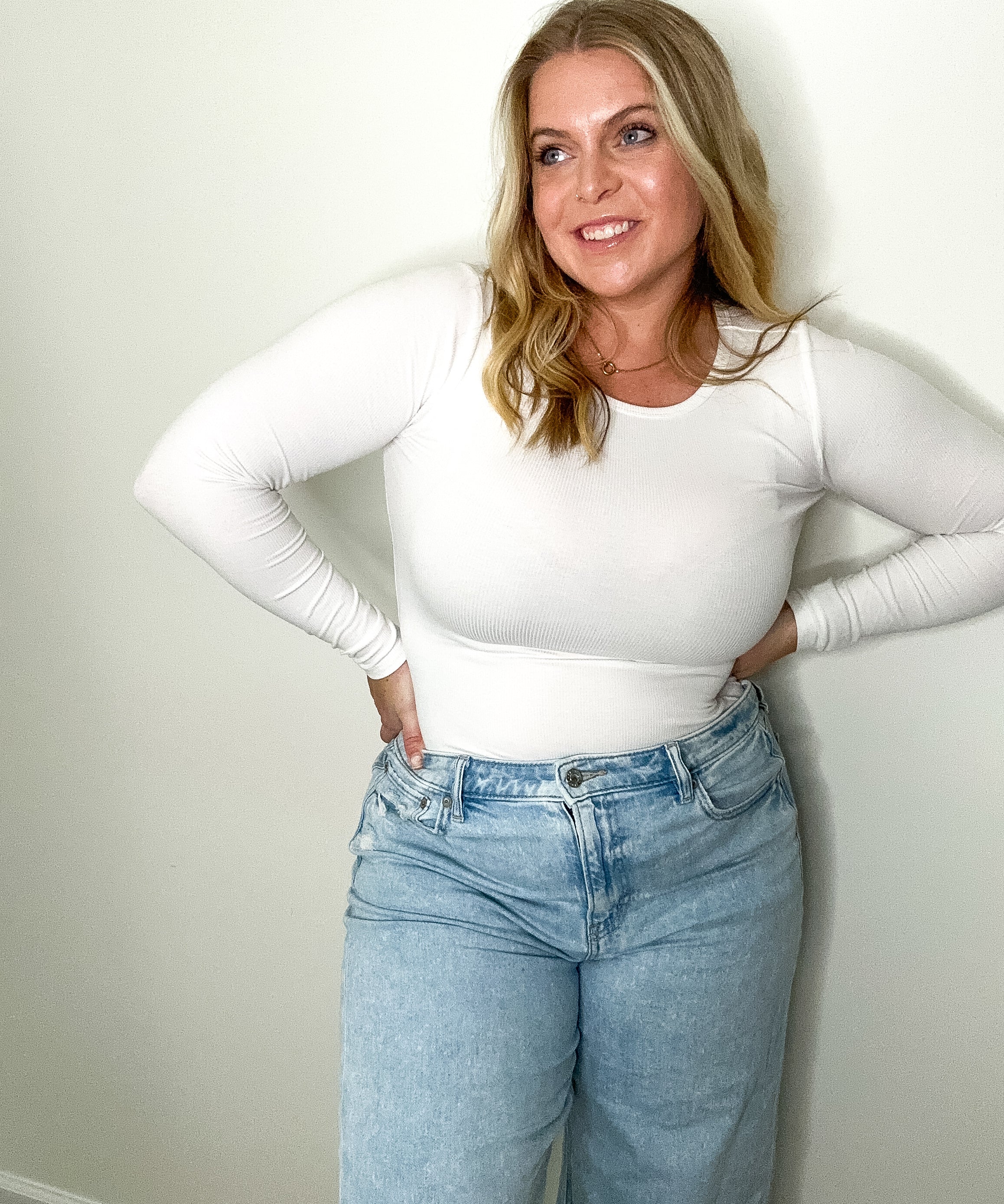 Woman wearing a white long-sleeve top and blue jeans against a plain wall.