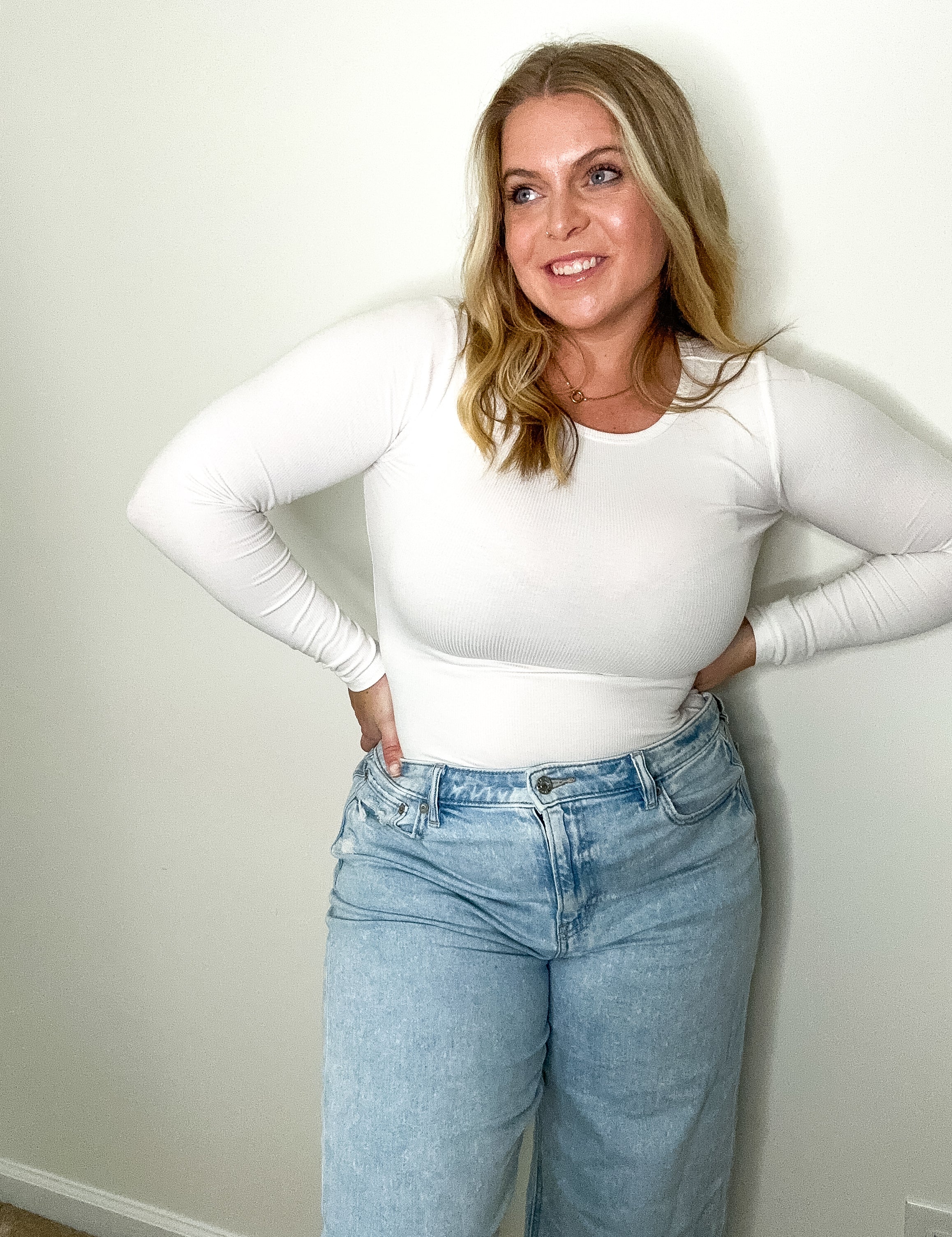 Woman wearing a white long-sleeve top and blue jeans against a plain wall.