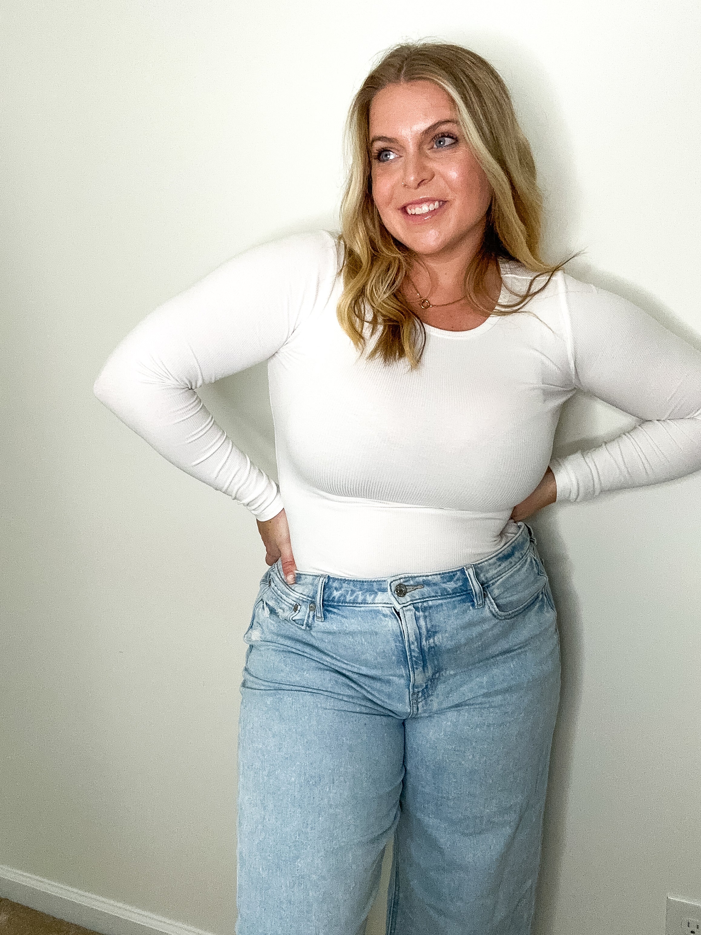 Woman wearing a white long-sleeve top and blue jeans against a plain wall.