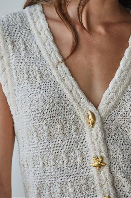 Woman wearing a textured white sleeveless top with gold buttons against a plain background