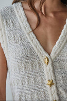 Woman wearing a textured white sleeveless top with gold buttons against a plain background