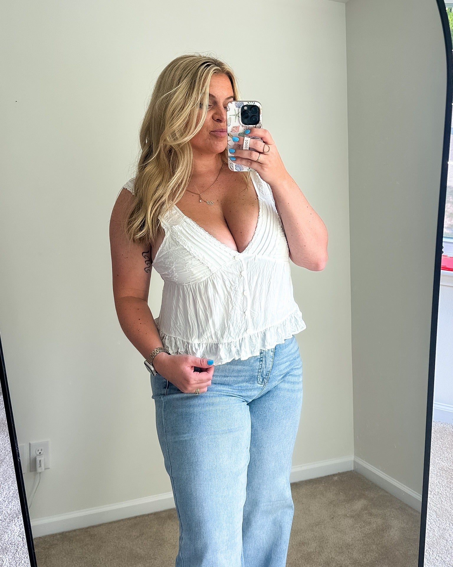 A woman takes a mirror selfie wearing a white sleeveless ruffled crop top and light blue jeans in a room with beige walls and carpet.