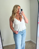 A woman takes a mirror selfie wearing a white sleeveless ruffled crop top and light blue jeans in a room with beige walls and carpet.