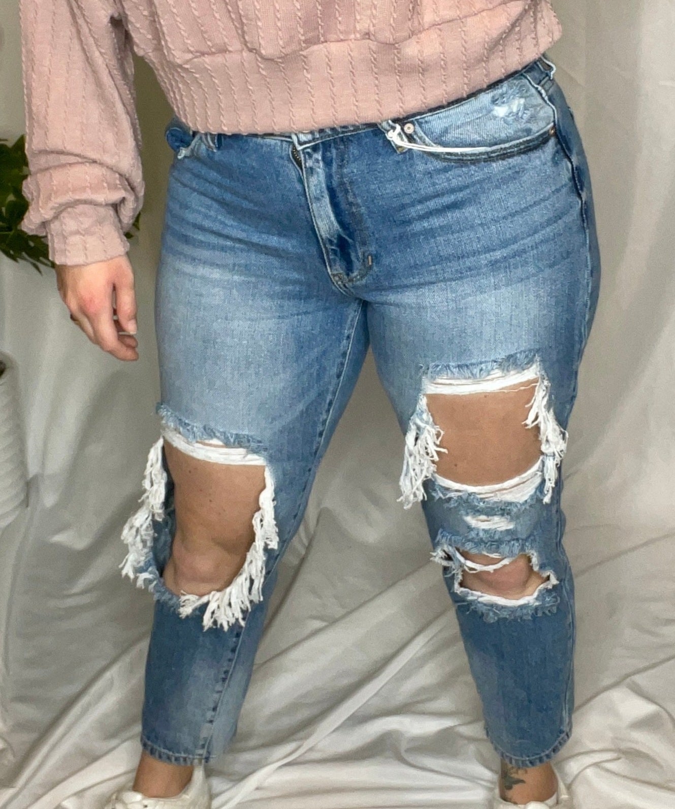 A person wearing a high-rise boyfriend fit jeans with large distressed rips, paired with a pink sweater and white shoes.