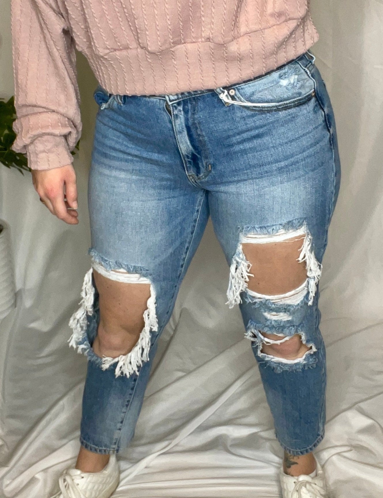 A person wearing a high-rise boyfriend fit jeans with large distressed rips, paired with a pink sweater and white shoes.