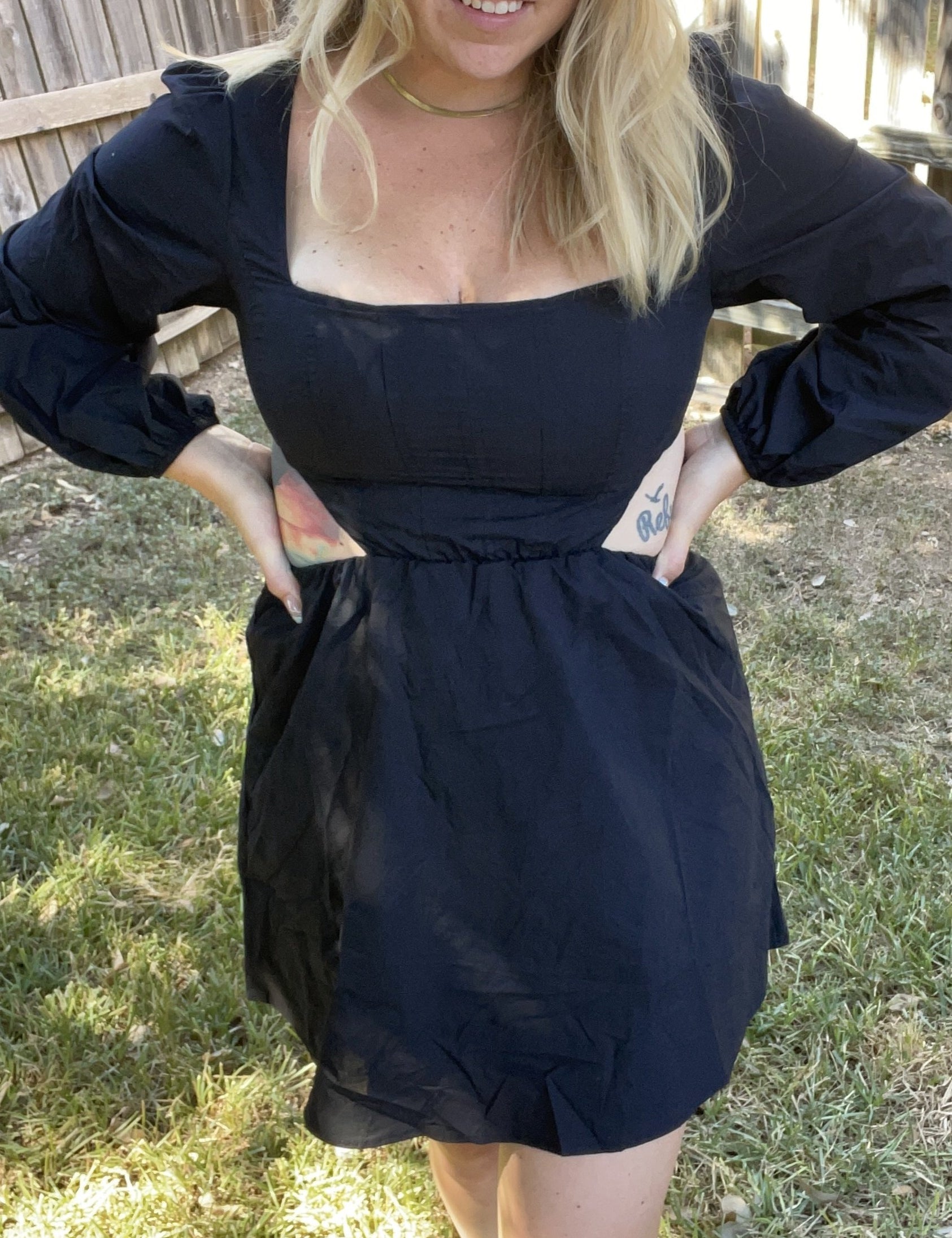 A woman wearing a black, short-sleeved, V-neck dress with a cut-out detail on the back, standing in an outdoor setting.